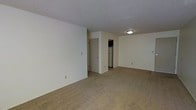 This single-floor space features an open layout with a spacious main living area finished in neutral, cream-colored walls that create a clean and understated atmosphere. The flooring throughout is covered in wall-to-wall beige carpet, adding a sense of warmth and continuity across the space. A textured popcorn ceiling with a centrally mounted flush light fixture provides straightforward overhead illumination. The living area is generously proportioned, offering flexibility for various furniture arrangements. Toward the rear of the space, a doorway leads into a compact kitchen area, where a refrigerator and what appears to be a range or microwave unit are visible, suggesting a practical and functional layout. A closet door is also visible near the kitchen entry, providing convenient storage. The overall design is simple and utilitarian, with minimal architectural embellishments. The neutral palette and open floor plan lend the space a versatile quality, making it easy to personalize. The single-floor layout ensures easy accessibility throughout, with no stairs to navigate between areas.