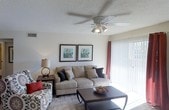 This single-floor residence centers around a well-appointed living room that establishes a warm, inviting atmosphere. The living room features neutral-toned walls paired with a textured ceiling equipped with a multi-blade ceiling fan with integrated lighting, ensuring both comfort and adequate illumination throughout the space. Natural light fills the room through a sliding glass door dressed with sheer white curtains and rich red accent drapes, hinting at outdoor access beyond.

The seating arrangement includes a beige upholstered sofa accented with decorative throw pillows in dark charcoal and tan tones, complemented by a boldly patterned accent chair featuring a medallion print with a red throw pillow. A dark-wood coffee table anchors the seating area, while a matching end table with a classic table lamp adds ambient lighting. Two matching framed botanical prints mounted side by side on the main wall provide a cohesive decorative focal point. The flooring transitions from carpet in the living area to tile near the sliding door. A hallway visible in the background suggests additional rooms beyond the living space.