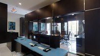 This single-floor office space showcases a contemporary design aesthetic defined by a striking contrast between rich, dark espresso cabinetry and cool light blue workstation surfaces. The main workspace features an elongated light blue desk equipped with multiple iMac computers, keyboards, and mouse pads, creating a clean and functional workstation setup. Floor-to-ceiling dark wood cabinetry lines the rear wall, incorporating recessed lighting and display niches that house decorative accents, including sculptural objects and abstract artwork, adding a polished, curated feel to the environment. A glass-walled conference room is visible beyond the main workspace, revealing ergonomic task chairs and a dark conference table. The glass partitions allow natural light to flow through the space while maintaining visual connectivity between areas. Large exterior windows within the conference room frame views of outdoor greenery, enhancing the sense of openness. The high-gloss tile flooring throughout reflects the ambient and recessed lighting, contributing to the bright, professional atmosphere. Overall, the space balances functionality with refined modern design, making it well-suited for a professional office or business environment.
