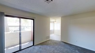 This single-floor residence offers a straightforward and functional layout designed for comfortable everyday living. The main living area features plush wall-to-wall carpeting in a neutral gray tone, providing a soft and inviting foundation throughout the space. A textured ceiling adds a classic architectural detail overhead, while a ceiling-mounted HVAC vent ensures year-round climate control.

A prominent sliding glass door opens directly to an outdoor balcony, allowing natural light to flow into the interior and offering a seamless connection to the outdoors. The balcony provides a private exterior space with views of surrounding greenery.

An open doorway leads into an adjoining room, which benefits from abundant natural light streaming through a large window dressed with vertical blinds. A ceiling fan is installed in this room, contributing to air circulation and adding a functional design element. Wall-mounted HVAC vents are present, further supporting a well-ventilated environment.

Throughout the space, clean neutral-toned walls create a bright and adaptable backdrop, making it easy to personalize with furnishings and décor. The overall ambiance is calm and understated, with practical features integrated throughout to support a comfortable and livable environment.