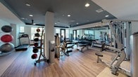 This fitness center offers a comprehensive workout environment designed to support a wide range of exercise routines. The space features warm hardwood-style flooring throughout, complemented by a dark, recessed ceiling with modern recessed lighting that creates a focused and energizing atmosphere. A teal accent band along the upper walls adds a stylish, contemporary touch.

The cardio zone is lined with Cybex treadmills, ellipticals, and stationary bikes, positioned along large windows that invite natural light and provide pleasant outdoor views of surrounding greenery. Full-length mirrors along one wall enhance the sense of space while allowing users to monitor their form during workouts.

The strength training area is well-appointed with a multi-station cable machine, adjustable benches, and a rack of dumbbells in varying weights. A dedicated functional training section includes a medicine ball rack and stability balls stored on a tiered stand. Towel service and storage shelves are conveniently accessible near the entrance.

Black-framed glass doors serve as the main entry, lending a sleek, modern aesthetic. A wall-mounted flat-screen TV adds entertainment options for those seeking a dynamic workout experience. The overall layout is open and well-organized, providing ample room to move freely between equipment stations.