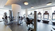 This fitness center presents a well-appointed exercise space with a sleek, modern aesthetic. The room is defined by its floor-to-ceiling windows along one wall, flooding the interior with natural light and offering expansive views of the surrounding urban skyline — an energizing backdrop for workouts. The ceiling features a refined paneled design with flush-mounted circular light fixtures that complement the room's contemporary style. Prominent cylindrical white columns add an architectural character to the space while subtly dividing workout zones. The flooring consists of durable dark grey tile, providing a practical and polished surface throughout. The facility is outfitted with a comprehensive selection of equipment, including multiple treadmills positioned near the windows, stationary bikes, and a range of Cybex-branded strength-training machines designed for targeted muscle work. The equipment is arranged to allow comfortable circulation between stations. The overall ambiance is clean, bright, and purposefully designed, making it a functional and visually appealing environment suited for a full range of fitness activities.