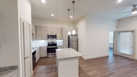 This single-story unit in Waxahachie, Texas presents a contemporary design with a neutral, light-toned palette and consistent wood-look flooring that flows throughout the living areas. The kitchen serves as a central highlight, featuring gray shaker-style cabinetry, a white subway tile backsplash, and granite countertops with subtle speckled detailing. A central island provides additional prep and seating space, illuminated by elegant pendant lights hanging from notably high ceilings. Stainless steel appliances, including a refrigerator, range, microwave, and dishwasher, complete the well-appointed kitchen. Open shelving and a built-in desk area offer added functionality nearby.

The bedroom features tall ceilings, a ceiling fan with light fixture, and a window fitted with blinds that allows natural light into the space. The bathroom continues the design cohesion with granite countertops, gray shaker cabinetry, a framed mirror, and tile flooring. A spacious walk-in closet with built-in shelving and hanging rods is accessible directly from the bathroom, offering generous storage. Throughout, the unit maintains a clean, modern aesthetic defined by consistent finishes, recessed lighting, and a well-organized layout.
