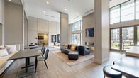 This contemporary amenity space is designed with an open, single-floor layout that maximizes both functionality and visual impact. Soaring double-height ceilings define the main lounge area, where floor-to-ceiling windows flood the interior with natural light and offer views of the surrounding exterior. The centerpiece of the lounge is a sleek, glass-enclosed linear fireplace topped by a wall-mounted flat-screen television, framed in light wood paneling and flanked by plush seating including a large sectional sofa and round ottoman stools. Wide-plank wood-look flooring runs throughout, unifying the various zones of the space. A dining and co-working area features modern tables and slatted chairs, while built-in shelving with accent lighting adds warmth and display functionality. The gourmet kitchen island showcases dramatic white marble-veined countertops, stainless steel appliances, and textured tile backsplash beneath soaring ceilings with a statement pendant light. The dedicated game room includes a foosball table, vintage arcade machine, dartboard, and a geometric chandelier, offering casual entertainment options. Throughout the space, curated large-scale artwork and carefully selected furnishings in neutral tones with rich accent colors contribute to a cohesive, upscale ambiance.