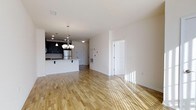 This single-floor space is defined by its open-concept layout, where the living area and kitchen flow together without interruption, creating a sense of spaciousness and ease of movement. Warm-toned hardwood flooring extends throughout, adding natural texture and brightness that is further enhanced by abundant natural light streaming across the floors.

The kitchen is a standout feature, anchored by dark navy cabinetry that provides a striking contrast against the crisp white walls and light countertops. A peninsula with seating overhang serves as both a functional workspace and a casual dining area. Stainless steel appliances, including a refrigerator, complement the modern aesthetic, while a subway tile backsplash adds a clean, classic detail. An elegant chandelier with multiple bulbs hangs above the peninsula, providing focused ambient lighting that enhances the kitchen's contemporary character.

The adjoining living area is open and versatile, offering a clean canvas framed by white walls and smooth ceilings. A paneled white door provides access to an additional room or storage space. Overall, the space presents a modern, well-lit, and functional environment with a cohesive design palette.
