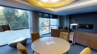 This single-floor space exudes a refined, contemporary aesthetic defined by a series of thoughtfully curated architectural and design elements. The most commanding feature is the ceiling, where a large circular recessed tray is finished in a warm, lustrous gold tone, housing soft overhead lighting that casts a welcoming glow across the room. The layered ceiling design adds depth and a sense of architectural distinction to the space.

At the center of the room sits a round light wood table accompanied by tufted accent chairs in a rich amber-gold upholstery, offering an inviting yet polished atmosphere suited for meetings or collaborative discussions. A lounge area in the background features neutral-toned seating, complemented by a sleek arc floor lamp with a brushed metallic finish, adding both function and visual interest.

The walls are finished in deep, cool-toned hues, contrasting elegantly with the warmth of the furnishings. Patterned sheer curtains frame expansive floor-to-ceiling windows that flood the space with natural light and provide open views of surrounding greenery and an outdoor terrace, enhancing the overall sense of openness and connection to the exterior environment.