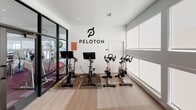 This single-floor fitness facility is thoughtfully designed to support a variety of workout routines in a clean, contemporary setting. The space is divided into distinct functional zones, beginning with a dedicated Peloton cycling studio. This enclosed room features multiple Peloton bikes with individual touchscreen monitors, separated from the rest of the facility by sleek black-framed glass partition walls and doors, creating a focused environment for cycling sessions. A large bank of frosted windows along one wall allows diffused natural light to fill the studio while maintaining privacy. The main workout area connects seamlessly through the glass-walled entrance, where a large wall-mounted flat-screen television and full-length mirrors enhance the training environment. The broader gym space provides room for additional equipment, including strength training machines and accessories. Light wood-toned flooring runs throughout, contributing to a bright and airy atmosphere, while recessed lighting ensures consistent illumination. The open layout and glass partitions create a sense of visual connectivity across the entire facility, making the space feel expansive and well-organized for a comprehensive fitness experience.