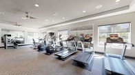 This fitness center offers a comprehensive, single-floor workout facility designed to accommodate a wide range of exercise routines. The space is thoughtfully organized into distinct zones, beginning with a dedicated cardio area lined with multiple treadmills, elliptical trainers, and stationary bikes, all positioned on protective floor mats beneath recessed lighting. Large windows along the perimeter flood the room with natural light and provide views of the surrounding outdoor environment, while roller shades offer adjustable privacy. Ceiling fans contribute to comfortable airflow throughout the space.

A strength training section features professional-grade cable machines, including Precor equipment, alongside weight benches and selectorized machines suited for a full-body workout. A separate free weights area includes a tiered dumbbell rack, a flat bench, stability balls, and a wall-mounted medicine ball rack, offering versatility for functional training. Large framed mirrors are positioned strategically throughout the facility, visually expanding the space and allowing users to monitor their form.

The neutral color palette of light gray walls, textured carpet flooring, and white ceilings creates a clean, modern ambiance. Wall-mounted televisions and a decorative art piece add finishing touches that enhance the overall environment. The facility also includes a ceiling-mounted fan in the tray ceiling section and direct access to an adjacent room, providing a well-rounded and professional fitness experience.