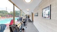 This single-level fitness center is thoughtfully designed to accommodate a wide range of exercise needs within a bright, welcoming environment. The space is divided into distinct functional zones, each outfitted with professional-grade equipment. A long, corridor-style cardio area runs alongside an expansive wall of floor-to-ceiling windows, offering unobstructed views of an outdoor swimming pool and surrounding greenery. This area houses elliptical trainers, stationary bikes, and additional cardio machines, all benefiting from abundant natural light that creates an energizing atmosphere. The main training hall features soaring ceilings accented with a teal upper wall, large mirrors that visually expand the space, and a lineup of treadmills positioned for an open, airy feel. Wall-mounted televisions provide entertainment during workouts. A dedicated strength training section is equipped with multiple resistance and weight machines featuring blue upholstered seating, complemented by organized shelving units stocked with dumbbells and kettlebells of varying weights. Exercise mats and balance equipment round out the functional training offerings. The flooring throughout is a durable, wood-look vinyl that ties the zones together cohesively. Textured concrete block walls and flush-mount ceiling lighting add to the clean, functional aesthetic of this comprehensive fitness facility.
