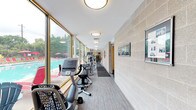 This single-level fitness center is thoughtfully designed to accommodate a wide range of exercise needs within a bright, welcoming environment. The space is divided into distinct functional zones, each outfitted with professional-grade equipment. A long, corridor-style cardio area runs alongside an expansive wall of floor-to-ceiling windows, offering unobstructed views of an outdoor swimming pool and surrounding greenery. This area houses elliptical trainers, stationary bikes, and additional cardio machines, all benefiting from abundant natural light that creates an energizing atmosphere. The main training hall features soaring ceilings accented with a teal upper wall, large mirrors that visually expand the space, and a lineup of treadmills positioned for an open, airy feel. Wall-mounted televisions provide entertainment during workouts. A dedicated strength training section is equipped with multiple resistance and weight machines featuring blue upholstered seating, complemented by organized shelving units stocked with dumbbells and kettlebells of varying weights. Exercise mats and balance equipment round out the functional training offerings. The flooring throughout is a durable, wood-look vinyl that ties the zones together cohesively. Textured concrete block walls and flush-mount ceiling lighting add to the clean, functional aesthetic of this comprehensive fitness facility.