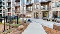 Located at 430 Englewood Ave SE in Atlanta's Grant Park area, The Upton's outdoor courtyard is a thoughtfully designed community amenity space set within a modern multi-story residential complex. The courtyard is framed on all sides by the building's contemporary facade, which blends brick masonry, dark metal accents, and expansive balconies to create a striking architectural backdrop.

At the heart of the space lies a generous artificial turf lawn, branded with The Upton's logo and outfitted with freestanding hammock loungers and cornhole boards for recreation. Adjacent to the lawn, a concrete patio hosts multiple distinct gathering zones: a pergola-covered seating area with string lights overhead, a fire pit surrounded by comfortable lounge furniture, and an outdoor grilling and dining station complete with umbrella-shaded tables and chairs.

Landscaped beds featuring ornamental grasses, mulched plantings, and young trees add natural texture throughout, while pathway lighting and a decorative stone water feature enhance the ambiance. A wrought iron fence encloses a separate pool area visible in the background, extending the range of outdoor amenities available to residents. The open-sky setting ensures abundant natural light throughout the day, making this courtyard a vibrant and functional outdoor living environment.