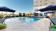Located at 4312 Yancey Rd in Charlotte, NC, this outdoor amenity space is designed to deliver a resort-style experience within a contemporary urban residential development. At its centerpiece is a generously sized swimming pool with crystal-clear blue water, framed by a wide stone-tiled deck that provides ample room for relaxation and recreation. Rows of sleek chaise lounges line the pool's perimeter, complemented by large navy blue market umbrellas that offer shaded retreats throughout the day.

The courtyard is enclosed by the development's multi-story buildings, which feature a sophisticated mix of brick, panel cladding, and large windows with private balconies overlooking the amenity below. The architecture creates a welcoming, semi-private atmosphere that balances open-air living with a sense of seclusion. Decorative pergola-style structures and private cabana areas add additional gathering spaces for residents.

Thoughtfully designed landscaping weaves through the courtyard, incorporating ornamental grasses, flowering shrubs, young trees, and raised planting beds that add texture, color, and natural beauty to the hardscaped environment. Stepped transitions with stainless steel railings connect varying levels of the outdoor space, while paved walkways guide movement throughout. The overall ambiance is polished, modern, and inviting—an ideal outdoor retreat in the heart of Charlotte.