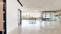 Located on W Olympic Blvd in Los Angeles, this contemporary leasing office spans a generously sized single floor, designed with a clean, modern aesthetic that balances functionality with refined style. Polished concrete floors run throughout the space, reflecting natural light that pours in through expansive floor-to-ceiling windows, offering open views of the surrounding Los Angeles streetscape. The layout is thoughtfully organized into distinct zones, including a welcoming lounge area furnished with plush seating — featuring a statement tufted blue sectional, curved cream armchairs, and marble-topped tables — creating an inviting environment for prospective residents. A café-style seating area with warm wood counter tables and light wood bar stools adds a casual, community-oriented atmosphere, complemented by framed artwork along the walls. Glass-enclosed private offices with black steel frames provide functional meeting spaces while maintaining visual openness. Natural greenery, including tall potted palms, adds warmth and vitality to the interior. Wood-paneled ceiling accents, linear LED lighting, and a dramatic large-format display wall showcasing architectural projects complete the sophisticated, gallery-like character of this professional leasing environment.