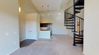 This two-story space makes an immediate impression with its dramatic vaulted ceilings that amplify the sense of volume and openness throughout the main living area. Soft, neutral-toned carpet flows across the generous open-concept floor plan, creating a warm and cohesive atmosphere. The kitchen is seamlessly integrated into the living space and features dark espresso cabinetry, stainless steel appliances, and a peninsula with an undermount sink — ideal for both everyday use and casual entertaining. Pendant lighting above the peninsula adds a refined touch, complementing the overall design. A set of double doors provides access to a closet or storage area, adding practical functionality to the layout. The standout architectural element is the elegant wrought iron spiral staircase positioned in the corner, which serves as both a functional connection to the upper floor and a striking visual focal point. Its dark metal finish contrasts beautifully against the light walls, reinforcing the space's modern aesthetic. The tall ceilings and open design come together to create an airy, loft-inspired atmosphere that balances style with livability across both levels.