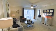 This single-floor residential space is thoughtfully designed to offer a comfortable and stylish living environment. The living area serves as the heart of the home, showcasing plush wall-to-wall carpeting that adds warmth underfoot. A ceiling fan with a brushed nickel finish sits centrally overhead, providing both function and a refined aesthetic touch. Crown molding runs along the perimeter of the ceiling, lending an air of elegance to the room.

Natural light floods the space through expansive sliding glass doors dressed with vertical blinds and floor-length drape panels in deep charcoal, creating a striking contrast against the neutral tones of the walls and flooring. The living area is generously proportioned, comfortably accommodating a full seating arrangement including a sofa, accent chair, and a sleek round coffee table with a mirrored base.

Decorative wall accents and carefully chosen furnishings contribute to the space's polished, contemporary ambiance. An open sightline connects the living room directly to the bedroom, which features its own window for additional natural light. The overall layout promotes an effortless flow between living spaces, making the home feel open, cohesive, and inviting throughout.