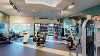 This fitness center offers a comprehensive, single-floor workout facility designed to accommodate a wide range of exercise needs. The space is organized into distinct zones, each purpose-built for different training styles. The cardio area features a row of treadmills positioned in front of large windows that invite natural light and provide outdoor views, complemented by a striking, large-scale fitness-themed mural that adds a bold visual identity to the room. Track lighting runs throughout the facility, ensuring bright, even illumination across all areas. The strength training section is well-appointed with a variety of resistance machines, including Instinct-branded equipment, along with free weights, medicine balls, battle ropes, and TRX suspension trainers neatly organized on dedicated Training Zone racks. A functional training area includes a cable machine and additional specialty equipment. Spin bikes with interactive screens offer an immersive cycling experience. The flooring throughout features durable rubber gym flooring with contrasting stripe accents, providing both safety and style. A tray ceiling with recessed lighting adds architectural definition to the main training floor. A reception counter, water fountain stations, and wall-mounted televisions round out the amenities, creating a fully equipped and visually engaging environment suited for diverse fitness routines.