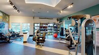 This fitness center offers a comprehensive, single-floor workout facility designed to accommodate a wide range of exercise needs. The space is organized into distinct zones, each purpose-built for different training styles. The cardio area features a row of treadmills positioned in front of large windows that invite natural light and provide outdoor views, complemented by a striking, large-scale fitness-themed mural that adds a bold visual identity to the room. Track lighting runs throughout the facility, ensuring bright, even illumination across all areas. The strength training section is well-appointed with a variety of resistance machines, including Instinct-branded equipment, along with free weights, medicine balls, battle ropes, and TRX suspension trainers neatly organized on dedicated Training Zone racks. A functional training area includes a cable machine and additional specialty equipment. Spin bikes with interactive screens offer an immersive cycling experience. The flooring throughout features durable rubber gym flooring with contrasting stripe accents, providing both safety and style. A tray ceiling with recessed lighting adds architectural definition to the main training floor. A reception counter, water fountain stations, and wall-mounted televisions round out the amenities, creating a fully equipped and visually engaging environment suited for diverse fitness routines.