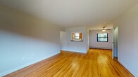 This single-floor residence offers a thoughtfully laid-out living space finished with warm, honey-toned hardwood flooring that flows consistently throughout the main living areas, bedrooms, and hallway. The open living and dining area is generously proportioned, featuring a ceiling fan and natural light streaming in through multiple windows that offer views of surrounding greenery. A distinctive pass-through window connects the living area to the kitchen, enhancing the sense of openness between the two spaces. The kitchen is equipped with rich wood cabinetry, dark granite-style countertops, a tile backsplash with a decorative mosaic accent strip, stainless steel appliances including a refrigerator and dishwasher, and a double sink beneath a window. The bedrooms are bright and well-sized, each featuring hardwood floors, baseboard heating, and windows that allow ample natural light. The bathroom is cleanly finished with large-format white wall tiles, a mosaic accent border, and a full soaking tub with a shower. A central hallway with overhead lighting connects the rooms efficiently. The space presents a clean, neutral palette throughout, making it highly adaptable to a variety of furnishing styles.