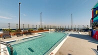 Located at 1035 Spring St NW in Atlanta's Midtown neighborhood, this rooftop amenity deck offers an exceptional outdoor retreat with striking views of the Atlanta skyline. The centerpiece is a generously sized swimming pool with in-water lounge chairs and a shallow sun shelf, all enclosed by sleek glass railings that preserve unobstructed sightlines to the city beyond. Surrounding the pool, a spacious sundeck is furnished with multiple chaise lounges arranged for both sunbathing and social gatherings. A vibrant, large-scale mural adds a bold artistic focal point to the lounge area, creating a dynamic and colorful ambiance. The outdoor kitchen features professional-grade stainless steel grills set against a clean, modern backdrop with floor-to-ceiling glass and steel-framed doors leading to interior amenity spaces. A dedicated wood-deck terrace with a ping-pong table and high-top seating provides an additional recreational zone, all framed by panoramic skyline views including iconic Midtown Atlanta towers. The overall design blends contemporary architecture with resort-inspired leisure, offering a well-rounded outdoor environment ideal for relaxation and entertainment.