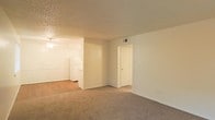 This single-floor space features an open-concept layout that connects the carpeted living area to the kitchen, creating a functional and cohesive floor plan. The living room is finished with wall-to-wall beige carpeting, neutral-toned walls, and a textured popcorn ceiling. A door off the main living area provides access to an additional room, likely a closet or utility space. The kitchen, visible from the living area, transitions to hard flooring and features white appliances, including a full-size refrigerator. A ceiling fan with a light fixture serves the kitchen area, contributing to ventilation and ambient lighting. The space maintains a clean, minimalist aesthetic with simple white doors, baseboards, and standard wall outlets. Ventilation is supported by ceiling vents throughout. The overall ambiance is straightforward and practical, offering a neutral canvas well-suited for a variety of furnishing and décor styles.