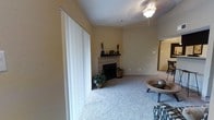 This single-floor residence offers a welcoming and functional layout designed for comfortable everyday living. The living room serves as the heart of the home, featuring plush wall-to-wall carpeting that adds warmth underfoot and a neutral, earthy color palette across the walls that creates a calm and inviting atmosphere. A standout feature of the space is the corner fireplace, elegantly framed with tile surround and topped with a dark wood mantle — an ideal focal point for the room. A ceiling fan with a light fixture is centrally positioned overhead, ensuring both comfort and practical illumination. Large vertical-blind-covered sliding glass doors span one wall, providing access to the outdoors and allowing ample natural light to fill the interior. The open layout connects seamlessly to adjoining areas, hinting at a spacious and well-considered floor plan. Subtle architectural details, including softly angled walls and clean transitions between rooms, contribute to the overall sense of ease and practicality. This single-story home presents a straightforward, move-in-ready living environment suited for those seeking comfort and simplicity.