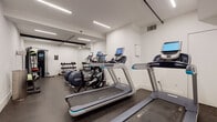 This fitness center is a thoughtfully designed, single-level workout space finished in a clean, contemporary style. The room is dressed in crisp white walls and durable rubber-speckled flooring, creating a hygienic and professional environment suited for focused exercise. Linear LED lighting runs along the ceiling, providing bright, even illumination throughout the space.

The cardio section is well-stocked with multiple treadmills and Precor elliptical machines, each equipped with individual digital display screens for performance tracking. A stationary cycle with an oversized interactive monitor adds a high-tech option for low-impact training. A full-length mirror along one wall visually expands the space while supporting proper form during workouts.

The strength and functional training area includes a multi-cable functional trainer with a digital interface, a tiered dumbbell rack holding a varied range of weights, and a medicine ball storage stand. Stability balls and additional accessories round out the functional fitness offerings. Built-in storage is provided via recessed cabinetry along one wall.

Overall, the fitness center balances a compact footprint with a comprehensive range of equipment, maintaining a clean, modern aesthetic that supports a focused and efficient workout experience.