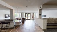 This single-floor community lounge is designed for comfort and social gathering, offering a well-appointed open-plan layout that seamlessly connects multiple functional zones. Large floor-to-ceiling windows and glass double doors flood the interior with natural light while providing views of the outdoor pool area and surrounding greenery, creating a bright and welcoming atmosphere throughout.

The main lounge area features a generous sectional sofa in rich dark leather, accented with decorative throw pillows in neutral and rose tones, arranged around a wooden coffee table atop a patterned area rug. A wall-mounted flat-screen TV adds a media-friendly dimension to the space. A second, more intimate seating nook is defined by built-in upholstered banquette seating with a bold damask print, complemented by a wall-mounted TV and a round white table with modern chairs — ideal for casual dining or small group gatherings.

The kitchenette is outfitted with warm wood cabinetry, dark stone countertops, a stainless steel sink, a beverage refrigerator, and a coffee station, all framed by a neutral mosaic tile backsplash. Stone tile flooring runs throughout the common areas, lending a cohesive, polished aesthetic. A reception-style counter near the entrance and pendant lighting complete the space's contemporary, community-focused design.