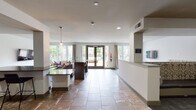 This single-floor community lounge is designed for comfort and social gathering, offering a well-appointed open-plan layout that seamlessly connects multiple functional zones. Large floor-to-ceiling windows and glass double doors flood the interior with natural light while providing views of the outdoor pool area and surrounding greenery, creating a bright and welcoming atmosphere throughout.

The main lounge area features a generous sectional sofa in rich dark leather, accented with decorative throw pillows in neutral and rose tones, arranged around a wooden coffee table atop a patterned area rug. A wall-mounted flat-screen TV adds a media-friendly dimension to the space. A second, more intimate seating nook is defined by built-in upholstered banquette seating with a bold damask print, complemented by a wall-mounted TV and a round white table with modern chairs — ideal for casual dining or small group gatherings.

The kitchenette is outfitted with warm wood cabinetry, dark stone countertops, a stainless steel sink, a beverage refrigerator, and a coffee station, all framed by a neutral mosaic tile backsplash. Stone tile flooring runs throughout the common areas, lending a cohesive, polished aesthetic. A reception-style counter near the entrance and pendant lighting complete the space's contemporary, community-focused design.