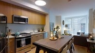 This well-appointed single-floor residence showcases a thoughtfully designed open-concept layout that seamlessly connects the kitchen, dining, and living areas. The kitchen is a standout feature, outfitted with warm wood cabinetry that extends to the ceiling, providing generous storage space. Stainless steel appliances — including a gas range, built-in microwave, and dishwasher — are integrated cleanly into the design, complemented by a sleek countertop and contemporary hardware. A large dark-toned dining table with matching chairs anchors the central space, creating a natural gathering point that bridges the kitchen and the living area beyond. The living area is bathed in natural light streaming through a prominent window, lending an airy, welcoming ambiance to the space. A mustard-yellow accent chair and tasteful décor add warmth and personality to the room. Neutral wall tones and light flooring throughout contribute to a clean, cohesive aesthetic. The layout flows intuitively from one area to the next, making the space feel both functional and inviting. Recessed and flush-mount ceiling lighting ensures the interior remains bright and well-lit at all hours.