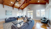 Located at 7029 W Friendly Ave in Greensboro, NC, this single-floor space offers a thoughtfully designed layout across a living room, office/game room, kitchen, and hallway. The living room is the centerpiece of the space, showcasing dramatic vaulted ceilings clad in warm natural wood planks, supported by exposed timber beams that lend a rustic yet refined character. A floor-to-ceiling stacked stone fireplace with a bold dark mantel serves as a striking focal point, complemented by recessed lighting, ceiling fans, and large multi-pane windows that flood the interior with natural light and offer views of the surrounding greenery. Warm hardwood floors run throughout, unifying the open layout. The kitchen features gray shaker-style cabinetry, white quartz countertops, a hexagonal tile backsplash, and black appliances, with a pass-through window connecting it to the adjoining living area. The game room offers a spacious, versatile environment with exposed wood ceiling beams, white beadboard wainscoting, gray walls, a billiards table, wall-mounted television, and a built-in bar with seating — ideal for recreation and entertainment. Together, these spaces create an inviting, well-appointed interior with a warm, cohesive aesthetic throughout.