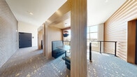 Located at 1875 Village West Pkwy in Kansas City, KS, this single-floor space offers a well-appointed layout comprising a hallway and dining room. The design blends warm natural materials with contemporary finishes throughout, creating a polished and inviting atmosphere.

The hallway serves as the connective core of the space, featuring striking slatted wood wall panels that add texture and visual warmth. An elevator and stairwell access are housed here, alongside patterned blue-and-gold carpet that continues throughout the floor. Vertical wood column dividers line the corridor, framing a built-in booth seating area with teal upholstered banquettes, a dark pendant light, and direct access to expansive windows overlooking the surrounding area.

The dining room is divided into distinct seating configurations. One area features a light wood table with integrated power access, surrounded by gray upholstered chairs beneath a sleek linear pendant fixture, complemented by a sideboard with a mirror. A second dining area is bathed in abundant natural light from floor-to-ceiling windows, offering open outdoor views. An exposed brick accent wall, curated artwork, and lounge-style seating further enrich the space, striking a balance between functional and aesthetically refined design.