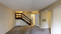 Located at 200 Carmen Avenue in Carrboro, North Carolina, this two-story residence offers a practical and comfortable layout across two levels. The main living area on the ground floor features plush carpeting and a prominent staircase with dark wood railings that serves as a central architectural element, leading to the upper level. Beneath the stairs, a convenient storage closet adds functional utility to the space. The kitchen is well-appointed with white cabinetry, gray speckled countertops, a stainless steel sink, and a classic subway tile backsplash, providing ample workspace and storage. An adjacent dining area transitions between the kitchen and living spaces, with wood-style flooring continuing the clean aesthetic. The bathroom features an extended vanity with dual sinks, gray countertops, white cabinetry, and a large mirror with bright overhead lighting. Upstairs, the carpeted bedrooms are generously sized, each equipped with ceiling fans and windows that allow natural light to filter in. One bedroom features dual sliding-door closets, offering substantial storage capacity. A private deck off the upper level overlooks a wooded setting of tall pine trees, providing a serene outdoor retreat.