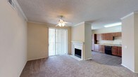 This single-story space showcases an open-concept layout that seamlessly connects the living area and kitchen. The living room is finished with wall-to-wall carpeting and features a white-mantled fireplace as a central focal point, adding warmth and character to the room. A ceiling fan with an integrated light fixture is centered overhead, contributing to both comfort and functionality. Large sliding glass doors fitted with vertical blinds allow natural light to fill the space while providing access to the outdoors. Crown molding accents the upper perimeter of the living area, lending a refined touch to the overall design. The textured ceiling adds visual depth throughout. Transitioning from the living room, the kitchen is finished with hard-surface flooring and features rich, medium-toned wood cabinetry that extends to the upper walls, offering ample storage. The kitchen is equipped with dark appliances and light-toned countertops, creating a practical and functional cooking space. The open sightline between the living room and kitchen enhances the sense of space, making the layout well-suited for everyday living and entertaining.