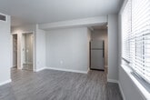 Located at 205 34th St. in Virginia Beach, Virginia, this single-floor unit offers a clean, modern aesthetic throughout. The main living area is open and airy, finished with durable wood-look plank flooring in a warm gray tone that extends seamlessly through the space. Neutral gray walls and white baseboards provide a contemporary backdrop, while a large window fitted with horizontal blinds allows natural light to fill the room. A ceiling-mounted vent and recessed lighting contribute to a comfortable, well-ventilated environment. The kitchen area is positioned along one wall and features gray cabinetry paired with a stainless steel refrigerator and a functional sink setup. The bathroom is a standout feature, clad in large-format marble-look porcelain tile that extends from floor to ceiling. It includes a gray vanity with a white countertop, brushed gold hardware, a wall-mounted mirror with overhead lighting, and a walk-in shower with matching tile surround. A dedicated walk-in closet with built-in wire shelving provides generous storage. The overall design emphasizes clean lines, neutral tones, and practical finishes, creating a fresh and move-in-ready living environment ideally situated near the Virginia Beach oceanfront.