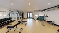 This fitness center is a thoughtfully designed, single-floor facility that provides a comprehensive workout environment. The space features a clean, open floor plan finished with durable light-toned flooring, crisp white walls, and dark baseboards that create a modern, polished aesthetic throughout.

The main workout area is generously proportioned, accommodating a wide variety of strength and cardio equipment. Notable pieces include a rowing machine, adjustable weight benches, a cable machine, a multi-function strength tower, and a full dumbbell rack with a neatly organized free weights selection. Floor-to-ceiling mirrors along one wall enhance the sense of space while serving a functional purpose for form monitoring during exercise.

Large windows and glass double doors flood the space with natural light and offer views of the surrounding exterior, creating an open, airy atmosphere. Recessed ceiling lighting provides consistent, even illumination throughout the room. The facility also features an interior railing along an open balcony-style section, adding an architectural dimension and allowing additional natural light to filter in from multiple angles.

Exposed ductwork along the ceiling adds an industrial touch to the otherwise clean, contemporary design. The double-door entry provides easy access, and the overall layout ensures ample circulation space between equipment, making for a comfortable and functional fitness environment.