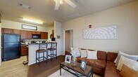 This efficiently designed 1-bedroom, 1-bathroom unit makes excellent use of its compact footprint across a single floor. The open-concept living area features warm-toned carpet, a ceiling fan with light fixture, and large windows dressed with bamboo shades and sheer curtains that allow generous natural light to fill the room. A sliding glass door provides additional brightness and outdoor access. The kitchen is equipped with rich cherry wood cabinetry, dark granite-style countertops, black appliances including a refrigerator, stove, and microwave, and a breakfast bar with seating for two — ideal for casual dining. A set of double doors in the kitchen conceals an in-unit laundry area, adding notable convenience. The bedroom is a comfortable retreat featuring plush carpet, a ceiling fan, a window with natural light, and enough space to accommodate a desk area for work or study. The bathroom is thoughtfully finished with a rich wood vanity, dark countertop, a single-basin sink, a framed mirror with overhead lighting, and wood-look flooring. Throughout the unit, neutral warm tones and consistent finishes create a cohesive and inviting ambiance suited to everyday living.