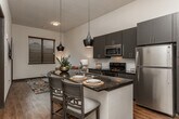 Located along Broad Street in Chattanooga, Tennessee, this single-floor unit showcases a contemporary design marked by bold contrasts and modern finishes throughout. The open-concept kitchen serves as the centerpiece, featuring dark espresso cabinetry, subway tile backsplash, and deep granite countertops that extend across a central island with bar seating. Stylish pendant lighting hangs above the island, adding an industrial-chic aesthetic to the space. Stainless steel appliances, including a gas range, built-in microwave, and full-sized refrigerator, complete the kitchen's functional appeal. Wood-style flooring runs through the main living and kitchen areas, contributing warmth and continuity, while vaulted ceilings in certain areas lend a sense of openness. The bedroom offers a quieter, carpeted retreat with natural light filtering through blinds-covered windows and ample space for furnishings. The bathroom maintains the same design language found throughout, with matching dark cabinetry, granite countertops, a large vanity mirror, and a full soaking tub with shower combination. Dark-framed interior doors and consistent hardware finishes create a cohesive, polished look from room to room. The overall ambiance is modern and refined, balancing bold design elements with comfortable livability.