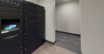 This single-floor package room is thoughtfully designed to provide a secure and organized space for parcel storage and retrieval. The room's most prominent feature is a tall bank of numbered black smart lockers lining one wall, offering multiple compartments of varying sizes to accommodate packages of different dimensions. The lockers' sleek, dark finish contrasts cleanly against the neutral, light-toned walls, creating a modern and functional aesthetic. The flooring transitions between durable stone-look tile in the main locker area and patterned carpet tile in the adjoining corridor, adding visual distinction between zones. Dark base molding runs along the perimeter of the walls, lending a polished, finished appearance throughout the space. The layout is open and accessible, with a wide corridor connecting the locker area to an adjacent room, ensuring ease of movement. The walls are finished in a warm greige tone, contributing to a clean and understated ambiance. Recessed lighting provides consistent, even illumination throughout. Overall, the space is well-appointed and purpose-built for efficiency, combining practical storage infrastructure with a clean, contemporary interior finish.