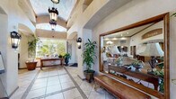 This single-story Mediterranean-inspired facility makes a strong first impression with a grand entry corridor showcasing soaring vaulted, barrel-arched ceilings, wrought-iron lantern pendant lights, and decorative wall sconces. Large-format tile flooring with an inlaid geometric border pattern runs the length of the hallway, flanked by lush potted tropical plants and a substantial framed mirror. Arched doorways connect the entry to a series of well-appointed interior spaces. The formal lounge features a white-surround fireplace, rich crown molding, tall arched windows dressed in dramatic deep-red drapes with ornate valances, and views of an outdoor pool. A carpeted multi-purpose room offers generous open floor space, recessed lighting, and direct access to the entry corridor and adjacent rooms. A dedicated screening room provides rows of leather theater-style seating, a wall-mounted flat-screen display, a built-in decorative fireplace, and an arched wrought-iron transom window. The kitchen is outfitted with warm wood cabinetry, granite countertops, stainless steel appliances, and tile flooring. A formal dining area features the same arched window design with floor-length red drapery. Throughout, textured plaster walls, consistent Mediterranean detailing, and abundant natural light create a cohesive, refined ambiance.