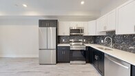 This thoughtfully designed single-floor space combines modern finishes with a clean, contemporary aesthetic throughout. The kitchen serves as a standout feature, showcasing a bold two-tone cabinetry scheme with dark lower cabinets and crisp white upper cabinets, all complemented by sleek white countertops and a striking dark marble-veined backsplash. A full suite of stainless steel appliances — including a French door refrigerator, electric range, over-the-range microwave, and dishwasher — adds both functionality and visual cohesion. Light wood-look flooring runs seamlessly through the kitchen and into the open living area, creating a bright and airy feel. The living space is generous and versatile, featuring recessed lighting, neutral toned walls, and a window that allows natural light to fill the room. The bathroom continues the marble theme with dark, veined floor tiles and a marble-surround soaking tub and shower combination fitted with a sliding glass door and matte black hardware accents. White paneled doors and clean white trim are consistent throughout, lending the space a polished, unified look. Overall, the unit presents a fresh, move-in-ready environment with high-quality finishes at every turn.