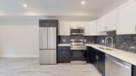 This thoughtfully designed single-floor space combines modern finishes with a clean, contemporary aesthetic throughout. The kitchen serves as a standout feature, showcasing a bold two-tone cabinetry scheme with dark lower cabinets and crisp white upper cabinets, all complemented by sleek white countertops and a striking dark marble-veined backsplash. A full suite of stainless steel appliances — including a French door refrigerator, electric range, over-the-range microwave, and dishwasher — adds both functionality and visual cohesion. Light wood-look flooring runs seamlessly through the kitchen and into the open living area, creating a bright and airy feel. The living space is generous and versatile, featuring recessed lighting, neutral toned walls, and a window that allows natural light to fill the room. The bathroom continues the marble theme with dark, veined floor tiles and a marble-surround soaking tub and shower combination fitted with a sliding glass door and matte black hardware accents. White paneled doors and clean white trim are consistent throughout, lending the space a polished, unified look. Overall, the unit presents a fresh, move-in-ready environment with high-quality finishes at every turn.
