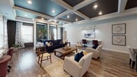 This well-appointed single-floor communal space offers a thoughtfully designed environment that blends comfort, functionality, and contemporary style. The open-concept layout connects several distinct zones seamlessly, all unified by rich hardwood-style flooring that runs throughout.

The lounge area serves as the centerpiece of the space, anchored by a deep navy blue sofa, plush neutral armchairs, and a wooden coffee table set atop a layered area rug. A pair of workstations with individual task lighting lines one wall, making the space equally suited for relaxation and productivity. Large windows invite generous natural light, framed by patterned drapery that adds a soft, decorative touch.

One of the most striking architectural features is the coffered ceiling, painted in a bold dark hue and accented with recessed lighting and white trim molding — a detail that adds depth and visual sophistication throughout the space. Structural columns punctuate the open floor plan, adding character without interrupting the flow between areas.

The kitchenette is outfitted with white shaker-style cabinetry, granite countertops, a dark tile backsplash, stainless steel appliances, and a sink — providing everything needed for casual refreshments. A dining table nearby offers a convenient spot for meals or informal gatherings.

A wide hallway connects additional rooms beyond the main area, maintaining the clean, modern aesthetic with consistent finishes and bright, airy tones throughout.