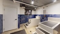 Located at 1415 Third Ave S in Birmingham, Alabama, this dedicated pet bathing facility is a compact, single-room space thoughtfully designed for self-service pet washing. The interior is defined by its striking bold blue geometric diamond-patterned tile wainscoting, which lines the walls at mid-height and gives the room a clean, modern character against the white upper walls.

The room houses two raised, white concrete bathing tubs, each elevated on stepped platforms with non-slip treads to allow easy pet access. The tubs are equipped with handheld spray fixtures and are positioned to allow comfortable use by attendants or pet owners. Between and around the tubs, anti-fatigue rubber matting covers the polished concrete floor, enhancing safety and comfort during use.

The ceiling features an exposed industrial aesthetic with visible ductwork and conduit running overhead, complemented by recessed and track lighting that provides bright, even illumination throughout the space. A wall-mounted mini-split unit ensures climate control year-round. A framed large-format photograph serves as a decorative accent on one wall, adding a touch of personality to the functional environment. Entry is provided through a glass-panel door. The overall design balances utility with a stylish, well-appointed aesthetic suited to a professional pet care setting.