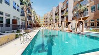 This expansive pool and grill area delivers a resort-caliber outdoor experience within a well-designed courtyard setting. The centerpiece is a large, crystal-clear swimming pool flanked by a series of elegant wood-framed cabanas positioned directly over the water. Each cabana features slatted canopy shades and plush orange-cushioned daybeds on warm wood decking, offering both shade and direct pool access. Additional lounge chairs are arranged along the stamped concrete pool deck, surrounded by mature palm trees that lend a tropical ambiance. A covered outdoor bar with stone-clad counters and bar seating provides a social gathering point beneath a wood slat pergola overhead. Adjacent to the bar, picnic-style dining tables offer casual al fresco seating. A striking natural stone fireplace serves as an architectural focal point in a dedicated lounge zone, surrounded by modern sectional sofas, woven armchairs, and accent tables—ideal for relaxed gatherings. The grill station features a built-in stainless steel gas grill set into a stone surround, accompanied by ample prep space. Native landscaping and ornamental grasses line the perimeter pathways, softening the hardscape and adding texture throughout. The entire space is framed by the surrounding multi-story building with balconied residences above.