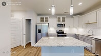 This single-story home in Wake Forest, NC, presents a clean, modern aesthetic defined by neutral tones, light hardwood-style flooring, and crisp white trim throughout. The interior maintains a cohesive and fresh feel across all rooms.

The kitchen serves as a focal point of the home, showcasing a large central island topped with granite countertops featuring subtle veining. White shaker-style cabinetry with glass-front uppers lines the walls, complemented by a classic white subway tile backsplash and stainless steel appliances, including a French door refrigerator, range, and microwave. Cylindrical pendant lights illuminate the island, adding a contemporary touch. An adjacent pantry with built-in shelving provides additional storage.

The living areas feature ceiling fans with integrated lighting and recessed lighting, contributing to a bright and comfortable atmosphere. Windows dressed with horizontal blinds allow natural light and views of surrounding greenery.

The bathroom includes a granite-topped vanity with white cabinetry and a soaking tub with tiled surround. A walk-in closet with wire shelving systems offers generous, organized storage. The dedicated laundry room includes a full-size washer and dryer, adding everyday convenience to this well-designed home.