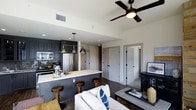 This single-floor space showcases an open-concept layout that integrates the kitchen and living area into one cohesive, functional environment. The kitchen is outfitted with dark, charcoal-toned cabinetry that contrasts elegantly with a light marble-style countertop along the island. A herringbone-patterned tile backsplash adds textural interest behind the stainless steel appliances, which include a full-size refrigerator and microwave. Three industrial-style pendant lights hang above the kitchen island, complementing the wooden bar stools that invite casual dining and conversation.

The living area flows naturally from the kitchen, anchored by a light-toned sofa accented with navy blue throw pillows. Dark hardwood flooring runs throughout, providing a warm and grounding contrast to the neutral walls. A ceiling fan with a matte black finish serves the living space, enhancing both aesthetics and airflow. Two white paneled doors in the background suggest access to private rooms or storage, maintaining a clean, organized feel. The overall ambiance is contemporary and inviting, balancing dark and light elements with modern finishes throughout.