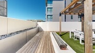 Located at 1031 N Park Ave in Tucson, Arizona, this rooftop amenity level at Malibu Tucson offers a thoughtfully designed outdoor retreat above the city. The space is anchored by a shimmering resort-style pool, complete with a sleek metal handrail entry and clean tile surrounds, perfectly positioned to capture Tucson's abundant sunshine and panoramic mountain views. Adjacent to the pool, a generously sized sun terrace is lined with rows of teak-framed chaise lounges set atop artificial turf, creating a vibrant and inviting poolside atmosphere. A second outdoor lounge area features warm wood decking, a built-in fire feature with stone accents, a pergola with natural timber beams, and comfortable seating, making it an ideal setting for relaxed gatherings. The upper rooftop level offers additional seating beneath a modern slatted shade structure, providing partial coverage while preserving sweeping views of the surrounding cityscape and distant mountain ranges. Access to the rooftop is provided through a contemporary interior corridor with polished concrete walls, wood-look flooring, and large glass doors that flood the transitional space with natural light. The overall design blends modern minimalism with warm natural materials, creating a cohesive and resort-inspired outdoor environment suited to Tucson's sunny climate.