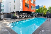 Located at 301 W Second St in the heart of downtown Santa Ana, California, this outdoor amenity space is a standout feature of a contemporary multi-story residential building. The architecture is defined by a striking combination of bold orange and clean white exterior panels, creating a vibrant, modern aesthetic that frames the pool deck on multiple sides.

The centerpiece of the space is a generously sized, resort-style swimming pool with crystal-clear blue water, accessible via integrated stainless steel handrails. Adjacent to the main pool is a separate spa or wading pool, set against a backdrop of mature trees that provide natural shade and a sense of seclusion. The surrounding pool deck is finished in large-format concrete pavers, offering a clean and durable surface throughout.

A well-appointed row of modern chaise lounge chairs lines one side of the pool, providing ample space for sunbathing and relaxation. Tall, square black planters filled with tropical and ornamental plants are thoughtfully placed around the deck, adding lush greenery and visual interest to the hardscape. Overhead, the open California sky allows for abundant natural light, enhancing the bright, airy atmosphere.

The space is enclosed by iron fencing and perimeter walls, ensuring privacy and security while maintaining an open, inviting feel. This outdoor amenity area offers a well-designed retreat ideal for year-round enjoyment in Southern California's favorable climate.