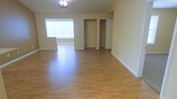 This single-floor space offers a clean, open layout designed for comfortable everyday living. The main living area is generously proportioned, featuring warm hardwood-style flooring that extends throughout, enhancing the sense of space and providing a cohesive aesthetic underfoot. Neutral, light-toned walls contribute to a bright and airy ambiance, complemented by a large window fitted with blinds that allows ample natural light to fill the room while offering privacy control. A ceiling fan with an integrated light fixture provides both air circulation and overhead lighting, making the space practical year-round. The layout includes a recessed closet alcove with a rod for hanging storage, as well as an additional door leading to a separate room or utility area, adding to the overall functionality of the space. Clean white baseboards frame the perimeter of the room, adding a crisp, finished look. The open floor plan provides flexibility for a variety of furniture arrangements and uses. Overall, the space presents a straightforward, well-maintained interior with practical features and a welcoming, understated character.
