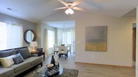 This well-appointed single-floor residence offers an open-concept design that connects the living and dining areas in a fluid, functional layout. The living room features vaulted ceilings accented by a classic ceiling fan with integrated lighting, adding both character and airflow to the space. Light hardwood-style flooring runs throughout, lending a warm and cohesive feel underfoot. Neutral gray walls provide a versatile backdrop, complemented by tasteful décor including a large abstract canvas artwork that adds a contemporary touch. The dining area sits just beyond the living space, bathed in natural light from multiple windows dressed with elegant white curtains. A pendant light fixture hangs above the dining table, creating an intimate atmosphere for meals. Large windows throughout the space invite generous amounts of natural light, enhancing the airy, open ambiance. The seamless flow between rooms makes the layout ideal for both everyday living and entertaining. Overall, the space strikes a balance between modern design and comfortable livability, with clean lines, thoughtful lighting, and a cohesive color palette throughout.